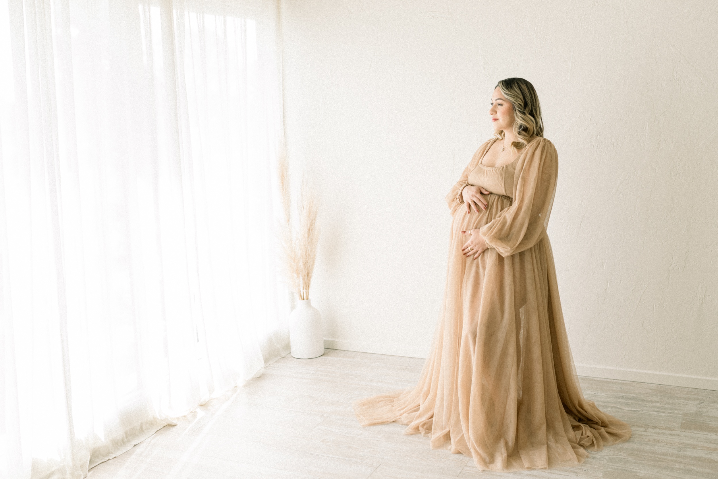 A smiling mother to be looks out a tall window while holding her bump in a flowing beige maternity gown after her 3d ultrasound in orange county
