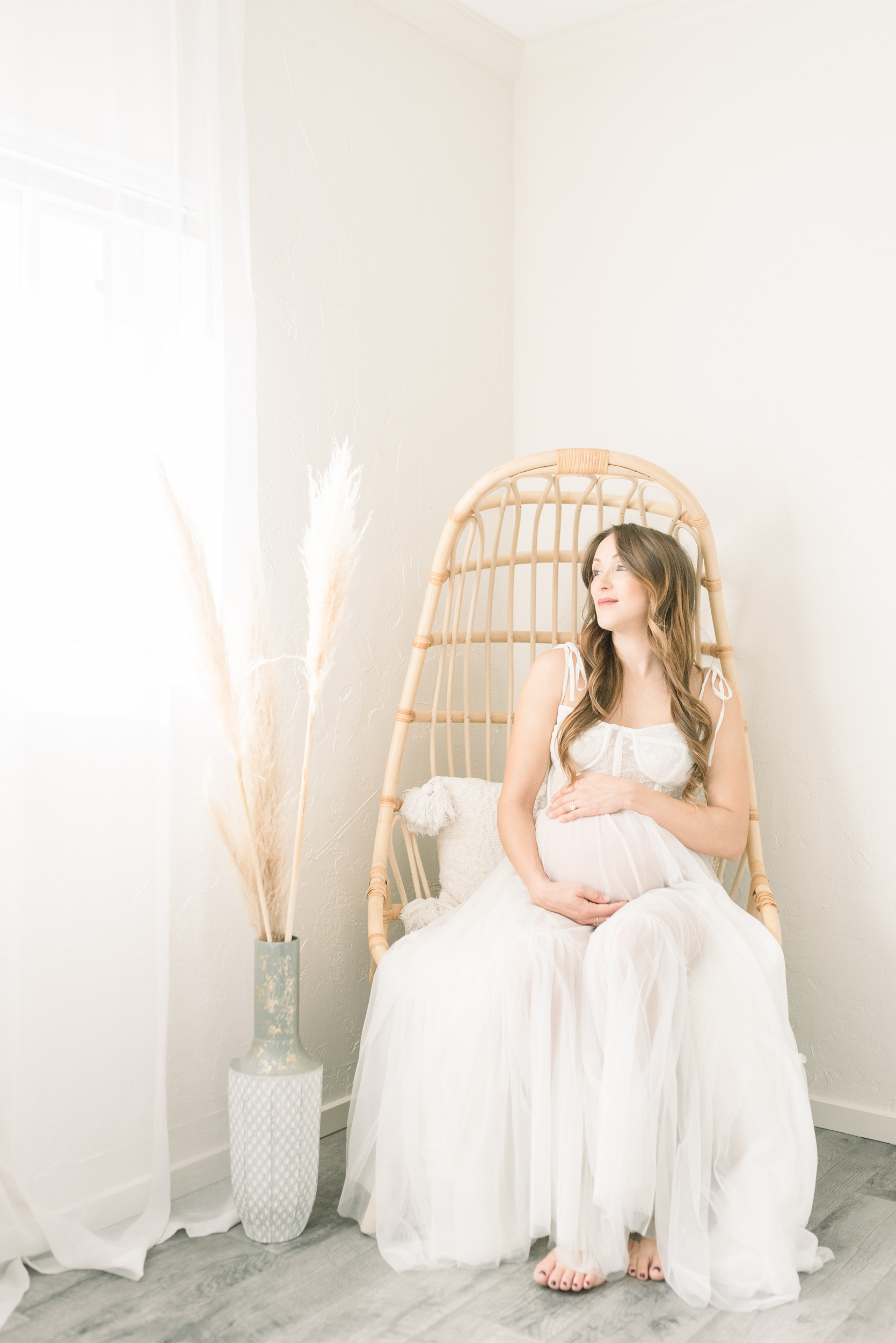 A smiling mother to be sits in a wicker chair in a corner of a studio holding her bump in a white gown