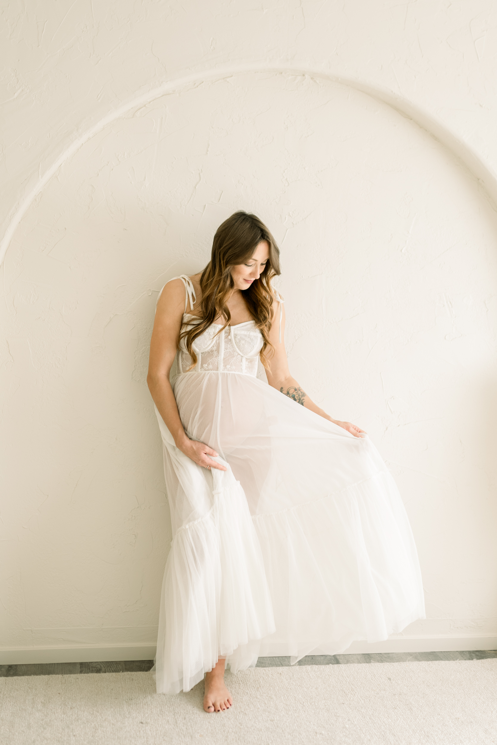 A pregnant woman leans on a plaster wall in an arch playing with her white maternity gown after visiting baby shower venues in orange county