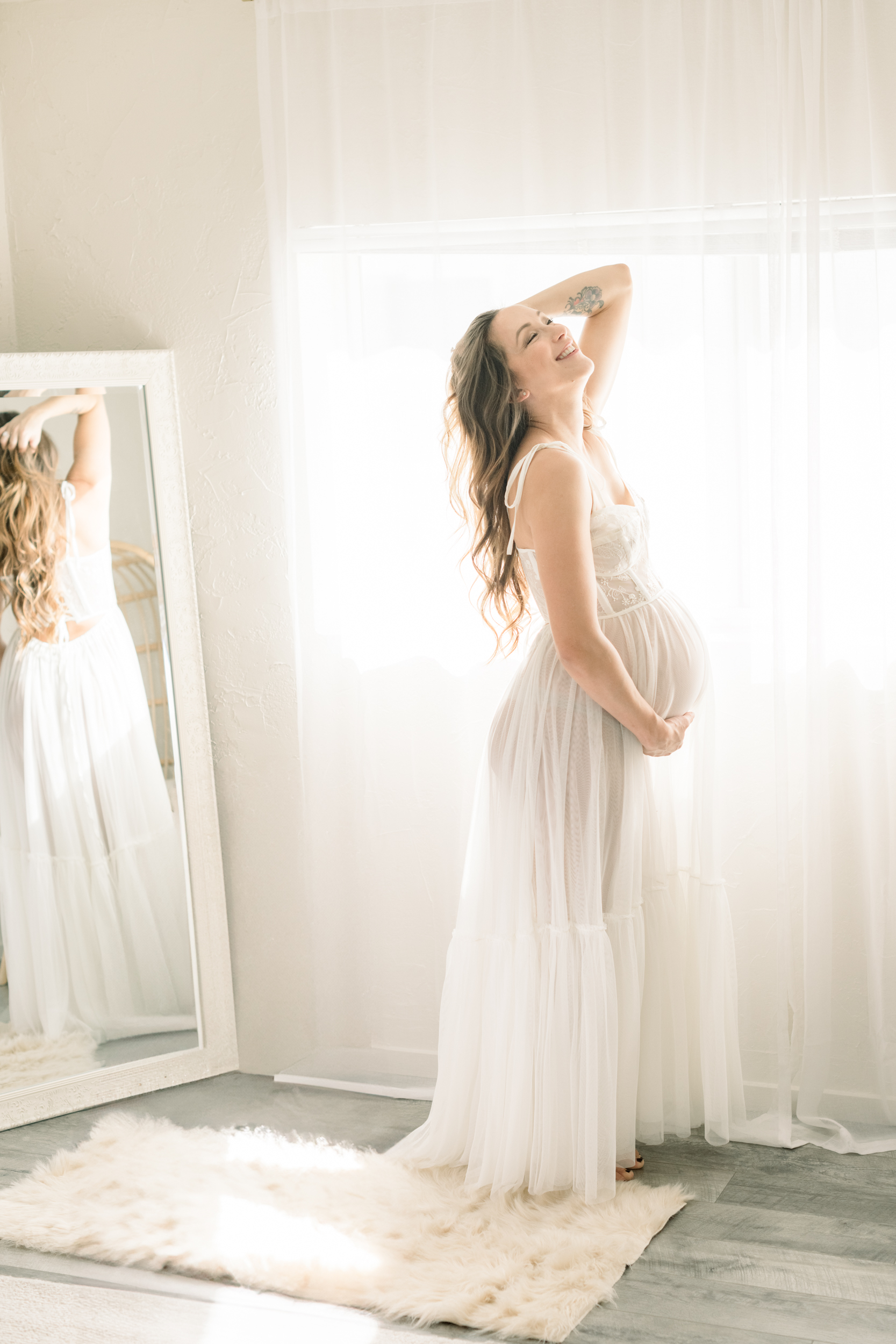 A smiling pregnant woman with a hand in her hair and other on her bump in a white maternity gown in a studio
