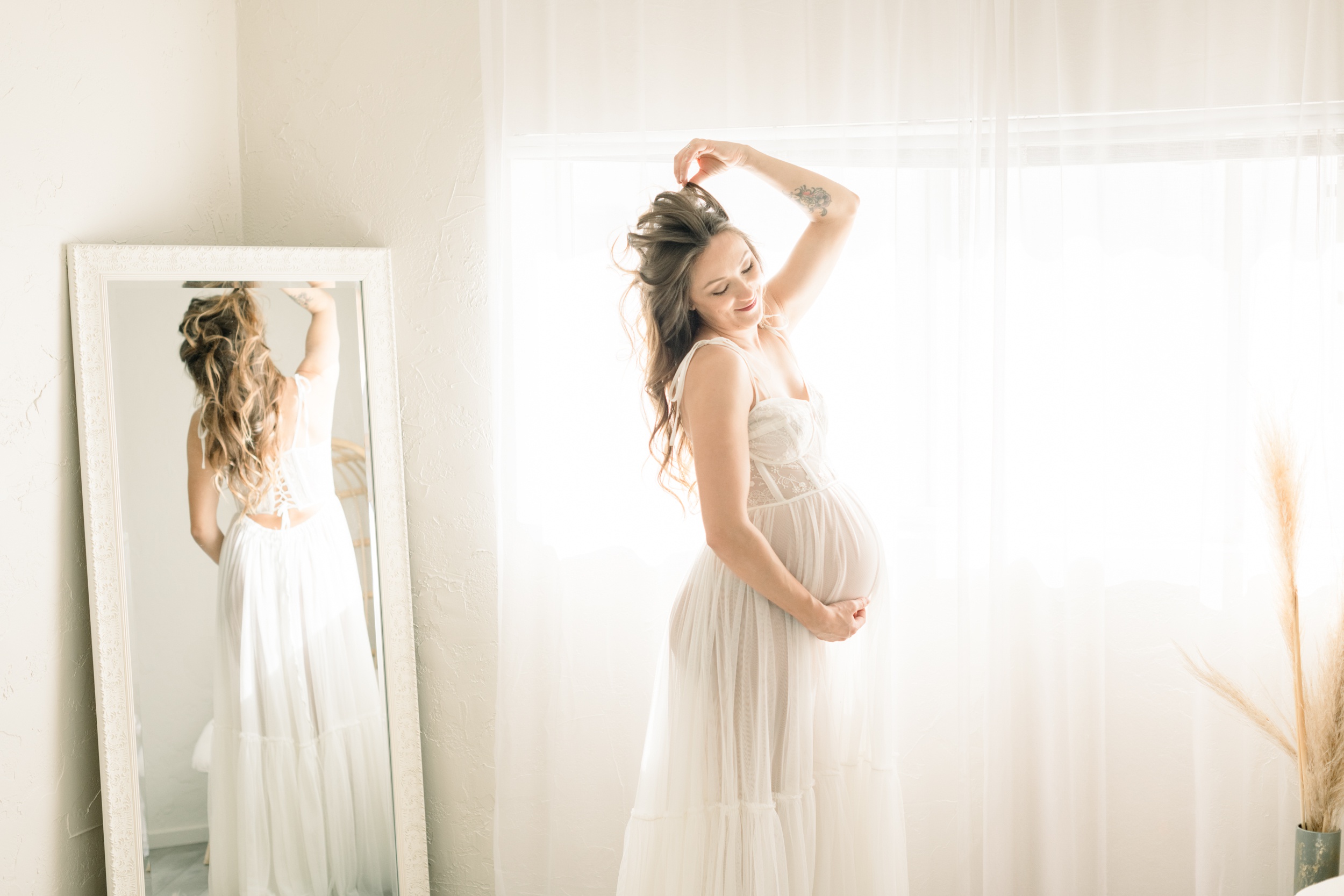 A happy mom to be stands in a studio playing with her hair in a white maternity gown after meeting doulas in orange county