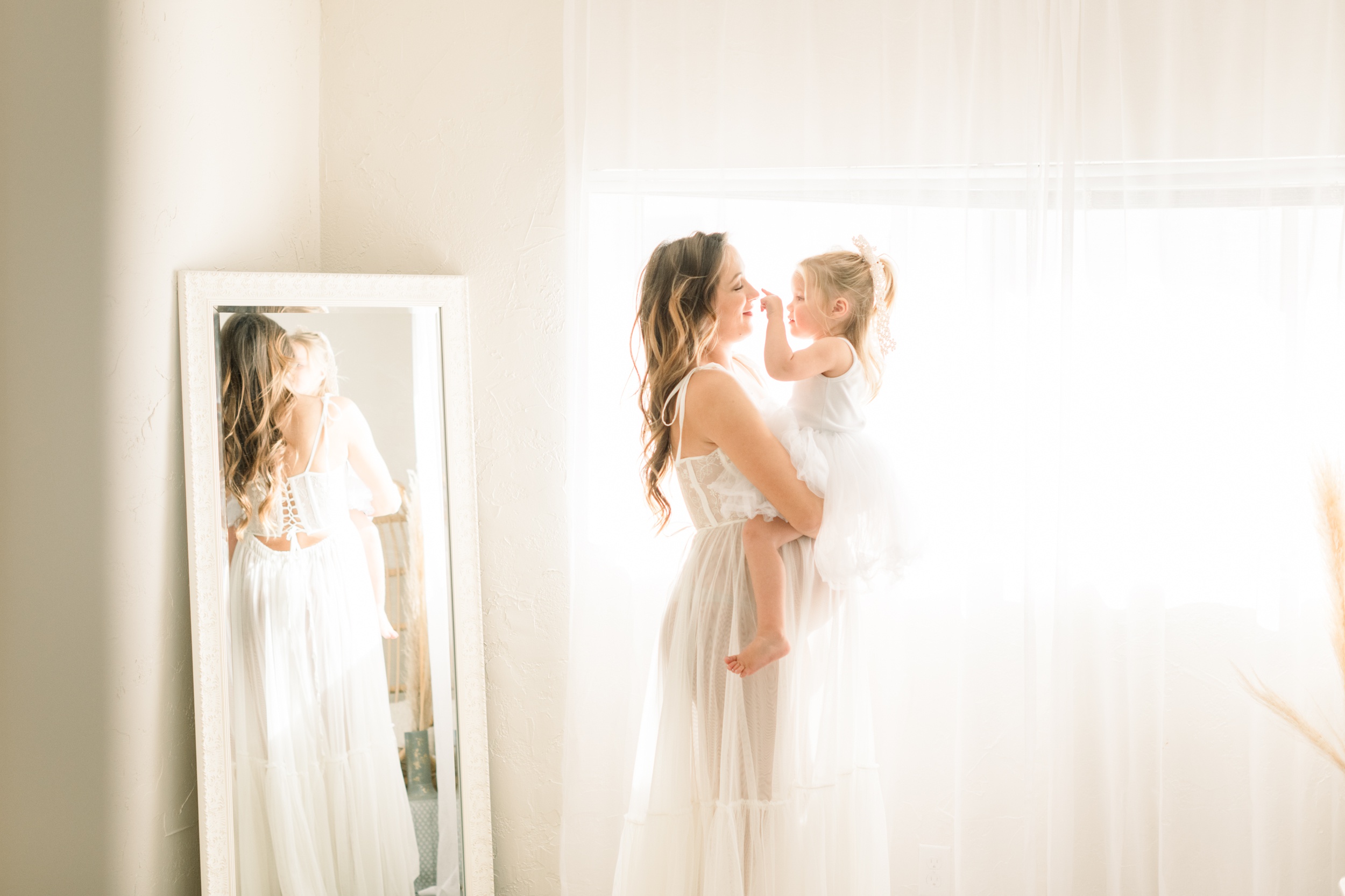 A happy pregnant mom plays with her toddler toddler daughter sitting on her bump both in white dresses after meeting doulas in orange county