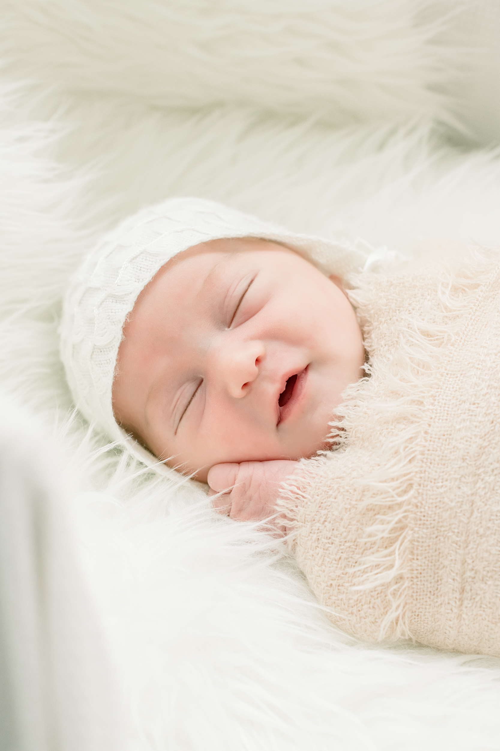 A closeup of a newborn baby sleeping in a white knit cap and beige blanket