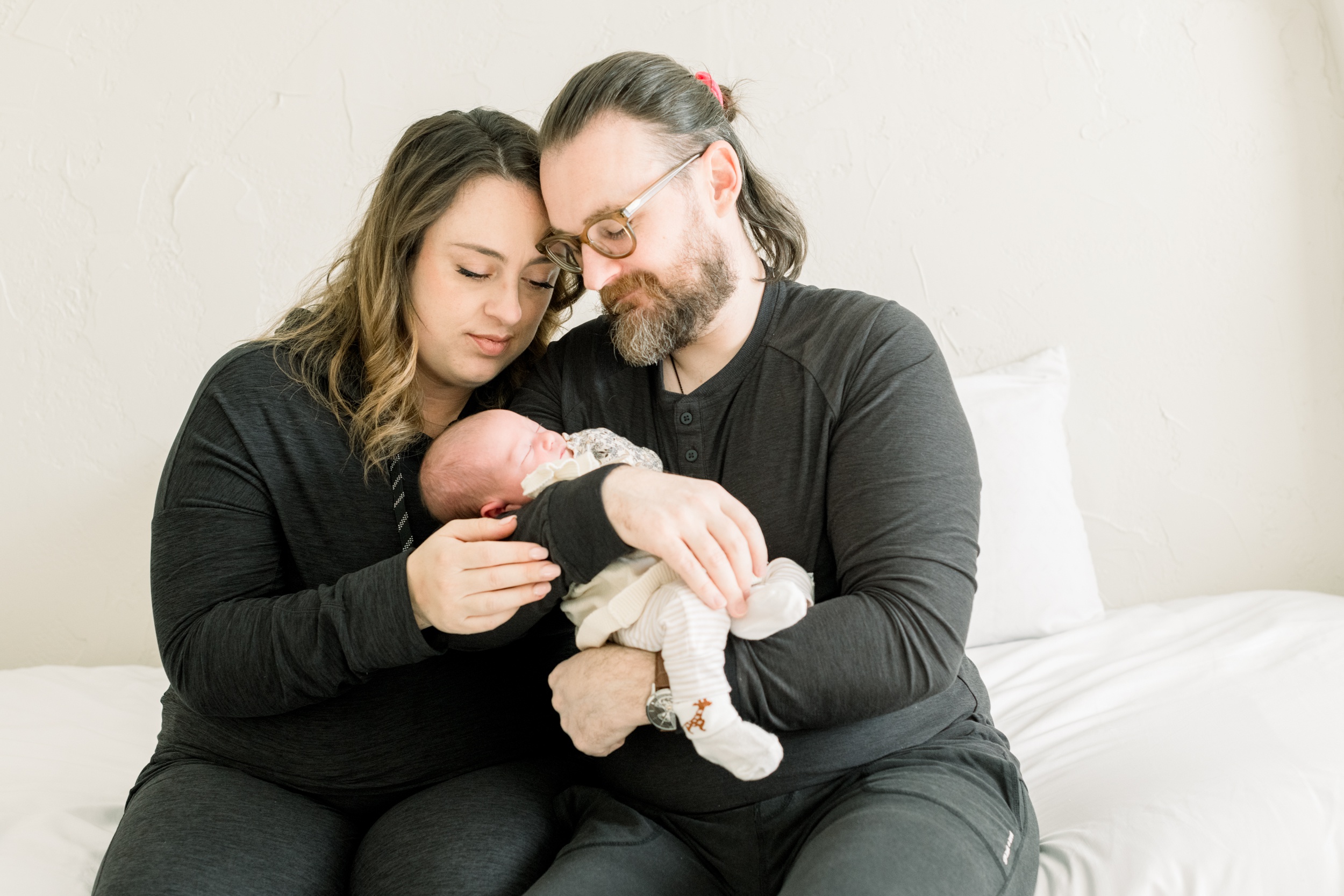 New mom and dad in matching dark shirts snuggle on a bed with their newborn in dad's arms after meeting nannies in orange county