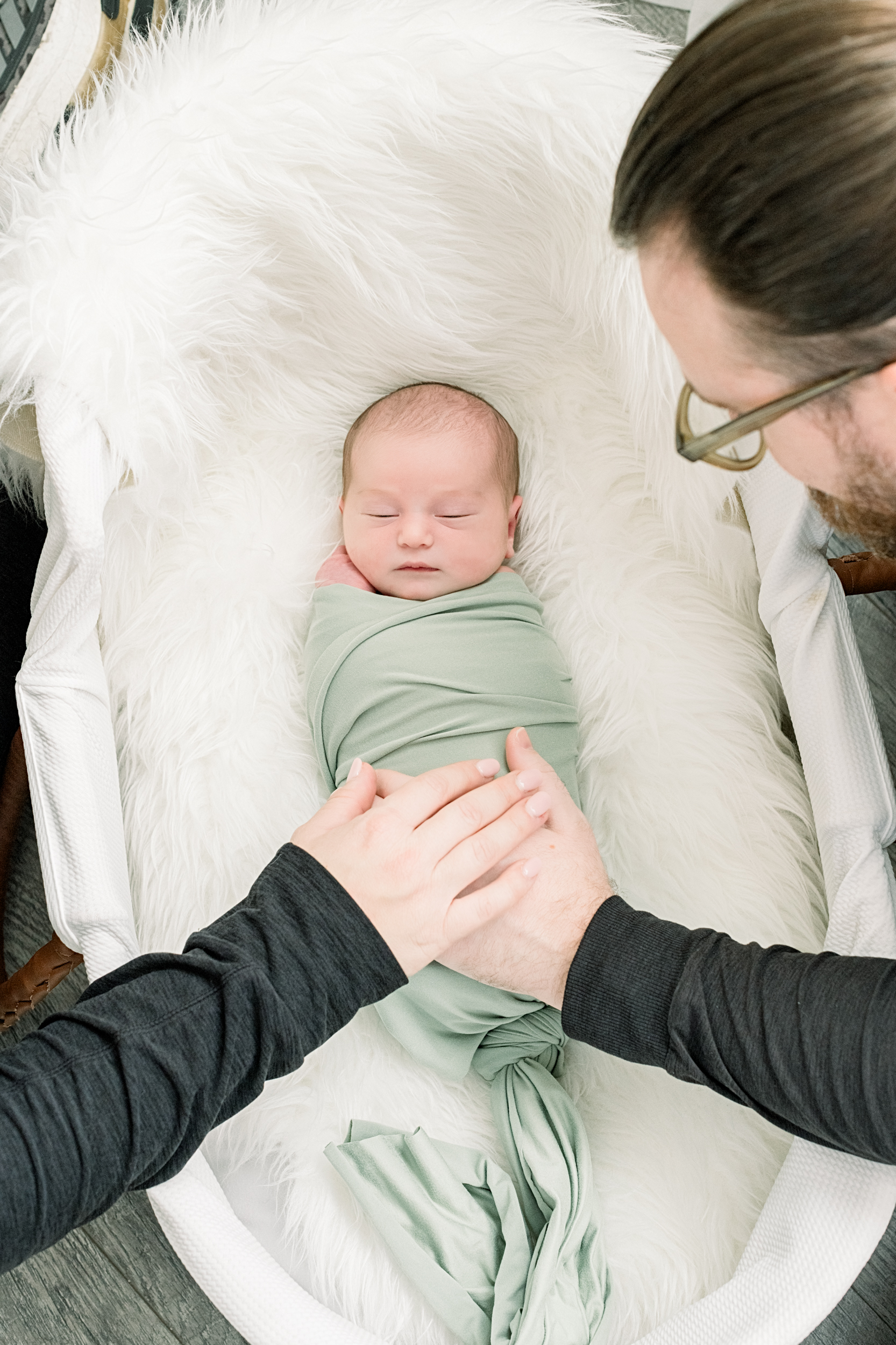 A newborn baby sleeps in a fur lined crib with mom and dad resting hands on it after meeting nannies in orange county