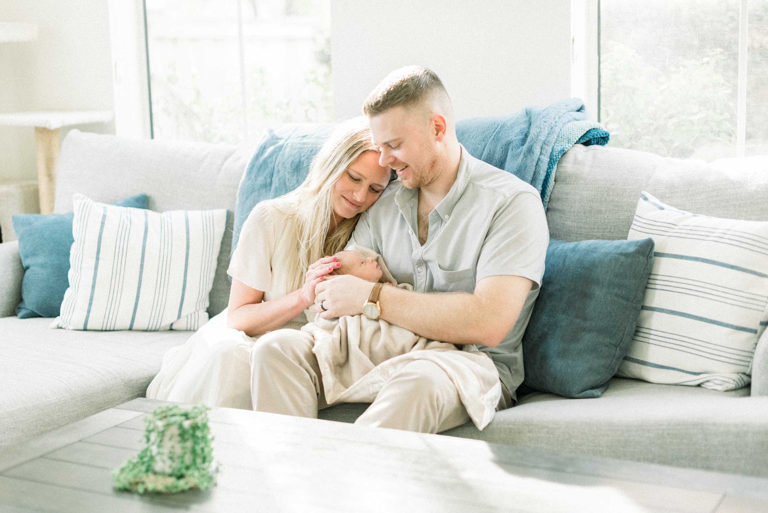 Happy new parents sit on a couch snuggling and admiring their newborn in dad's lap and arms before finding pediatric dentist in orange county