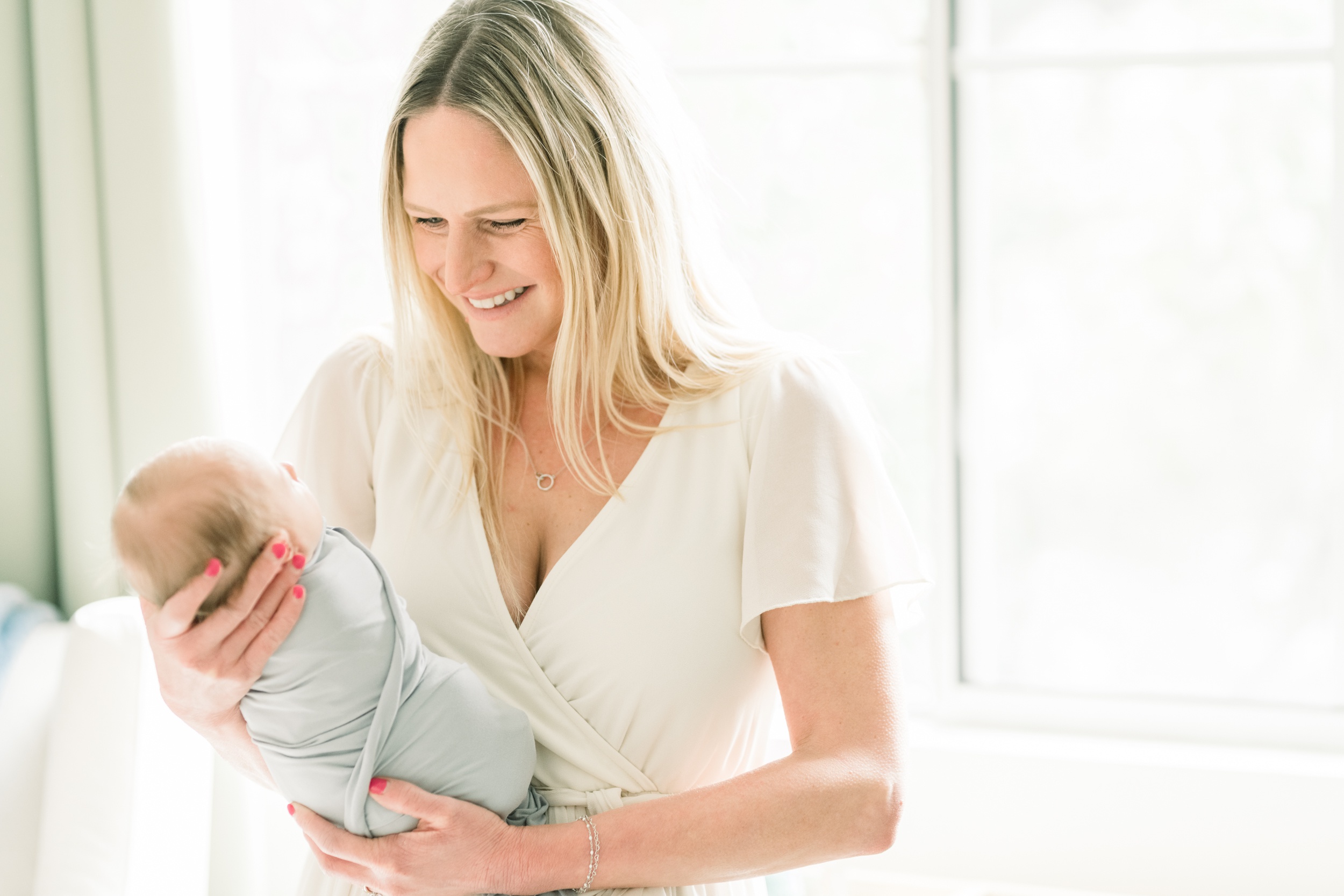 A giggling new mother in a white dress holds her sleeping newborn baby in her hands