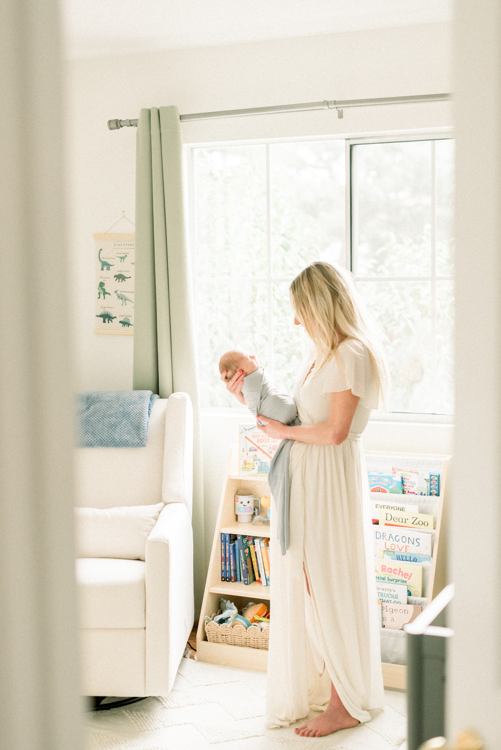 A new mother stands in a nursery holding her sleeping newborn in front of her in a white dress after some pelvic floor therapy in orange county