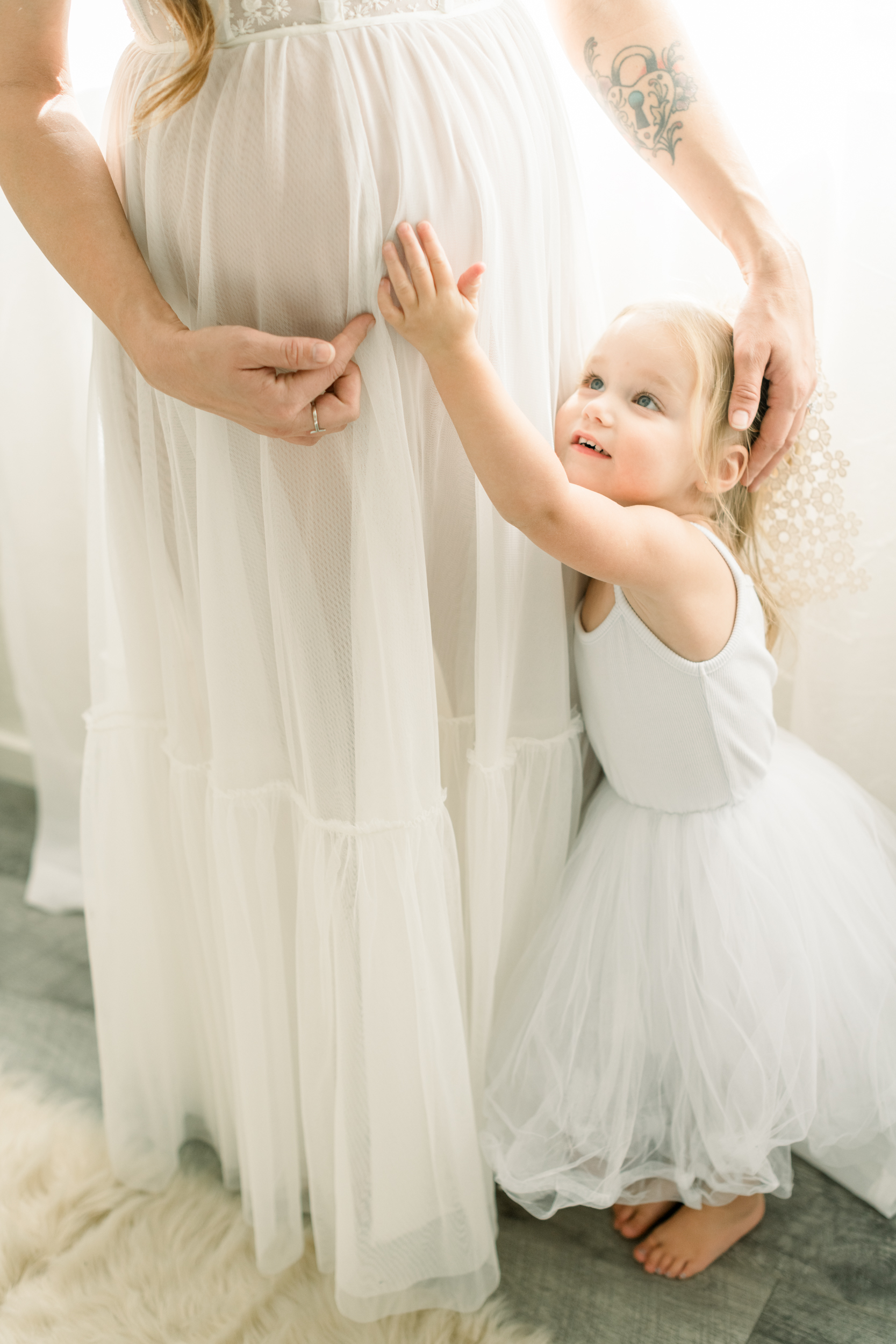 A toddler girl in a white dress reaches a hand up to mom's pregnant belly while standing in a studio after a prenatal massage in Orange County