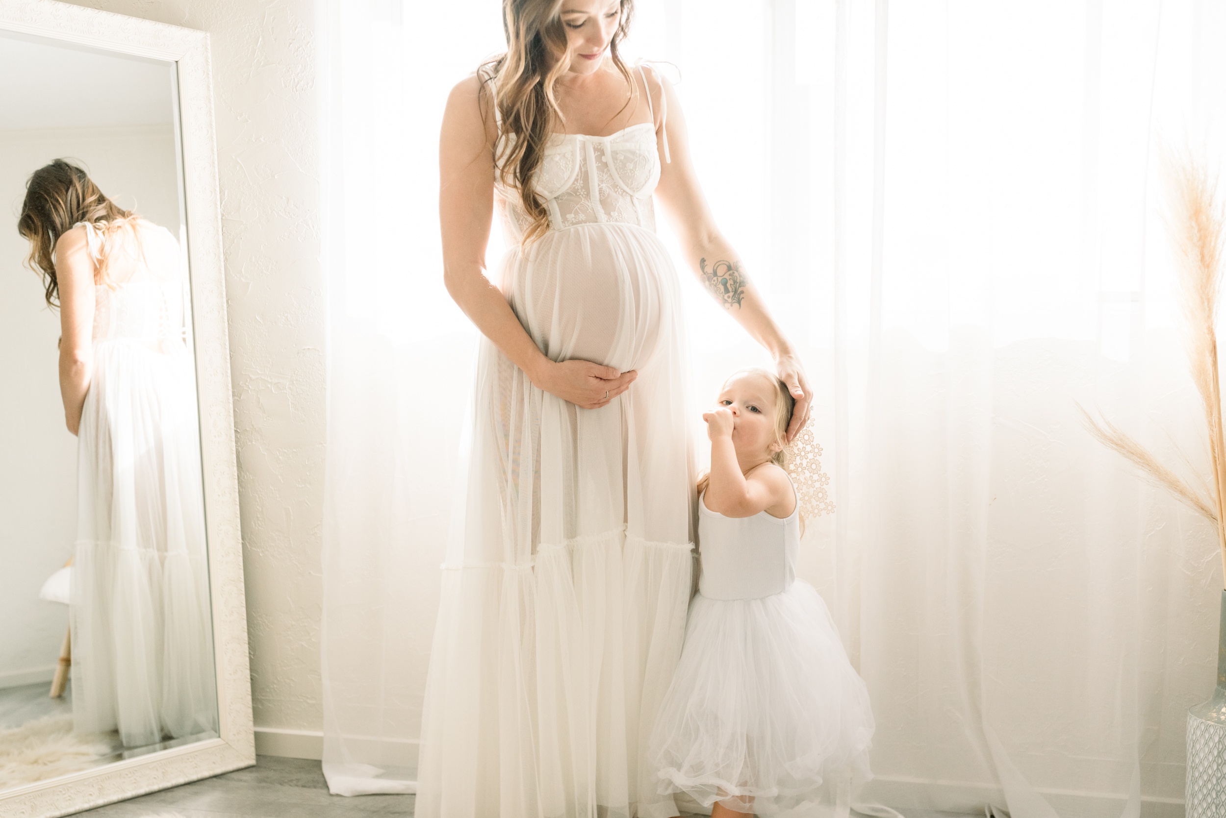 A pregnant mother stands in a white maternity dress with her toddler daughter at her side in a mirror with a thumb in her mouth after a prenatal massage in Orange County
