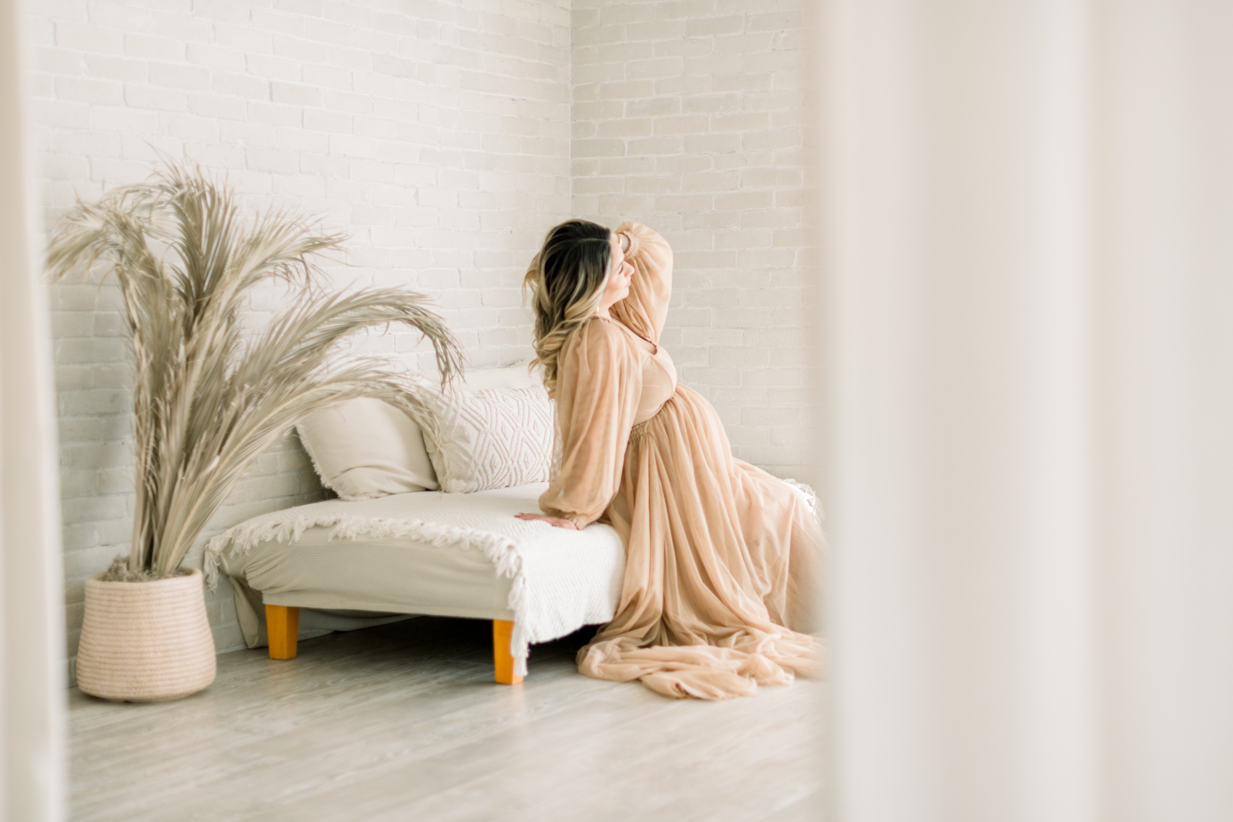 A pregnant woman leans back on a day bed in a studio with a hand in her hair in a beige maternity gown after some prenatal yoga in orange county