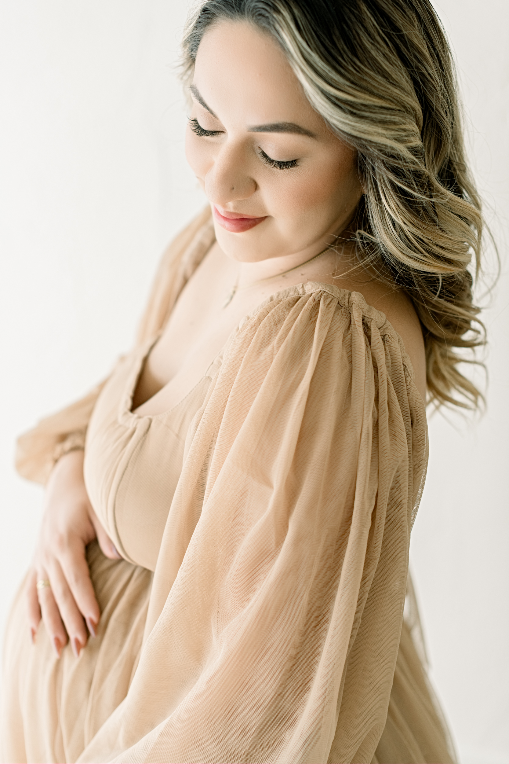 A mom to be in a beige maternity gown smiles down her shoulder with a hand resting on her bump