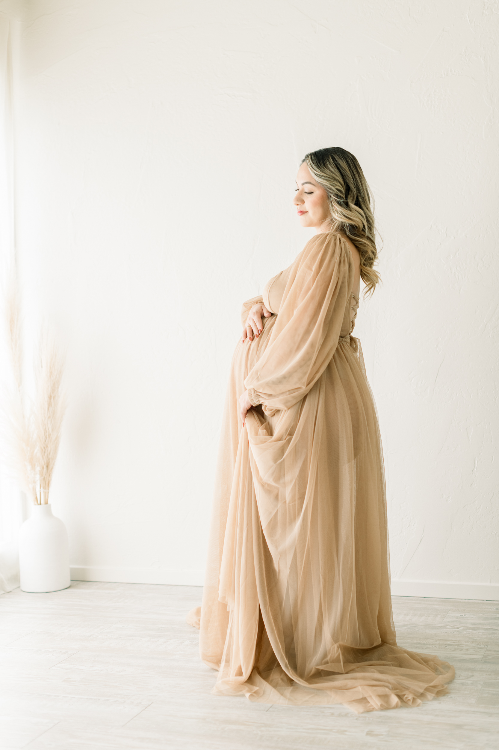 A smiling mother to be enjoys holding her bump in a beige maternity gown while standing in a white room after some prenatal yoga in orange county