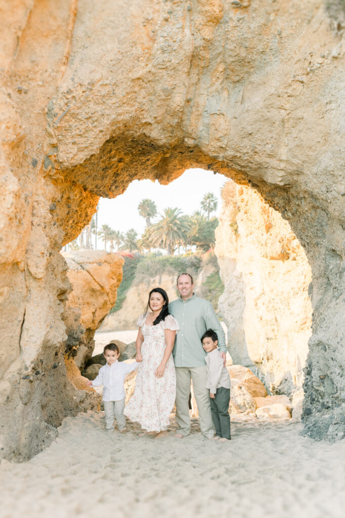 Laguna Beach family photographer, Amy Captures Love takes a photo of a family standing under a rock structure in Laguna Beach, California