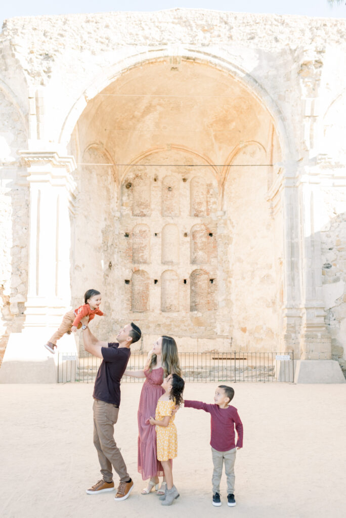 Family of 5 laughs while watching the youngest get tossed into the air during a fall family photoshoot at the Mission San Juan Capistrano with Amy Captures Love