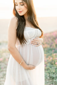 Woman glowing while she poses in a sheer white corset style reclamation dress along the floral beach bluffs in Huntington Beach California with Amy Captures Love