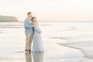 Couple stare out over the pacific ocean while in a calm embrace and they look stunning in their outfits as they shoot along beach for their maternity session taken by Amy Captures Love in Orange County California.