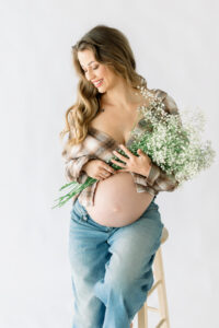 pregnant mama poses with her belly exposed while wearing a 90's flannel draped open and holding a baby's breath bouquet and wearing denim while shooting with Amy Captures Love in Huntington Beach, California.
