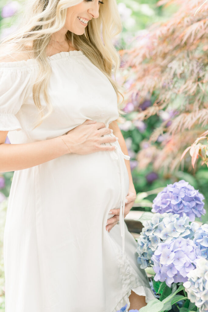Woman stands in garden next to flowers contemplating her maternity session date and when to schedule it
